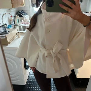 Vit cape-kappa med knytband - Stilren vit cape-kappa med hög krage, stora knappar och brett knytband i midjan. Kappan har vida ärmar och en mjuk, tjock känsla som ger en elegant siluett. Perfekt för dig som vill ha en trendig och unik look.