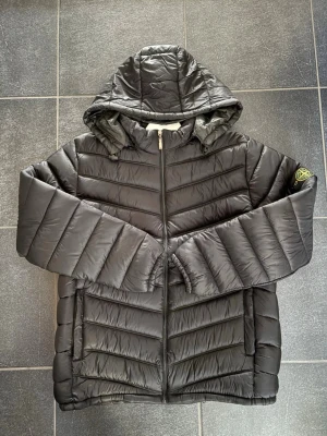 Svart Stone Island dunjacka - En clean svart dunjacka från Stone Island med huva och dragkedja framtill. Jackan har quiltad design, två sidofickor och klassisk logotyp på ärmen. Perfekt för kyliga dagar och enkel att matcha med olika outfits.