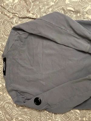 Grå sweatshirt från C.P. Company - Säljer en stilren grå sweatshirt från C.P. Company med rund hals och ribbade muddar. Tröjan har en ikonisk linsdetalj på ärmen och en ficka med dragkedja. Perfekt för dig som gillar streetwear och enkel design.