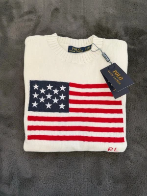Vit stickad tröja Polo Ralph Lauren USA flagga  - Vit stickad tröja från Polo Ralph Lauren med amerikansk flagga i rött, vitt och blått på bröstet. Rund halsringning och ribbstickade muddar. RL-broderi i rött nedtill. Klassisk och stilren design med tydlig preppy-vibe.