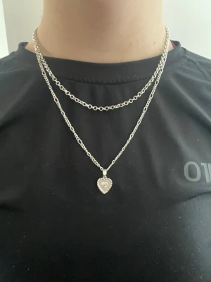 Silverfärgat halsband med hjärthänge - Säljer ett stilrent silverfärgat halsband med ett gnistrande hjärthänge dekorerat med små pärlor. Kedjan är medelgrov och ger en trendig look. Perfekt accessoar för att lyfta din outfit. Tveka inte på att höra av dig om du har några frågor! Svarar snabbt💕
