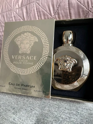 Versace Eros Pour Femme Eau de Parfum - Lyxig parfym från Versace, Eros Pour Femme, i en rund, spegelblank guldfärgad flaska med Medusa-huvud och dekorativa mönster. Flaskan rymmer 100 ml och kommer i en matchande exklusiv ask. Perfekt för dig som gillar eleganta och ikoniska dofter.