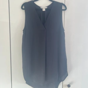 Svart ärmlös v-ringad topp - Snygg svart ärmlös topp med v-ringning och lätt omlott framtill. Toppen har en lös passform och är tillverkad i ett mjukt, tunt material som ger ett elegant fall. Perfekt att styla med jeans eller kjol för en trendig look.