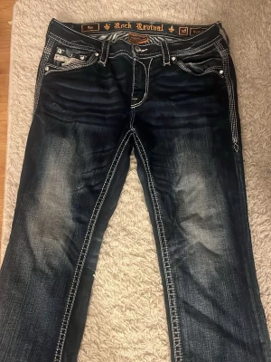 Rock Revival mörkblå jeans - Säljer ett par mörkblå jeans från Rock Revival med tydliga kontrastsömmar och slitningar på benen. Jeansen har klassisk femficksdesign, detaljerade fickor och en snygg tvättad look. Perfekta för dig som gillar streetwear och vill sticka ut.