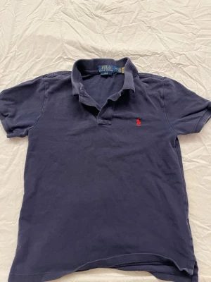 Mörkblå piké från Polo Ralph Lauren - Klassisk mörkblå pikétröja från Polo Ralph Lauren med röd broderad logga på bröstet. Slim fit-modell med korta ärmar och mjuk krage. Perfekt till jeans eller shorts för en clean och tidlös look. OBS barnstorlek