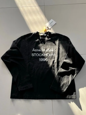 Svart långärmad topp Acne Studios - Svart långärmad topp från Acne Studios med tryck 'Acne Studios STOCKHOLM 1996' i vitt framtill. Klassisk rund halsringning och avslappnad passform. Tillverkad i mjuk bomull, perfekt för en stilren och enkel look.