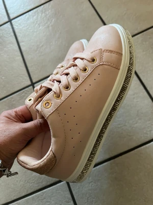 Ljusrosa sneakers med gulddetaljer - Snygga ljusrosa sneakers med guldfärgade öljetter och glittrig gulddetalj på hälen. Skorna har vita sulor med en glittrig kant och är gjorda i ett slätt material. Perfekta för dig som vill ha en clean och trendig look.
