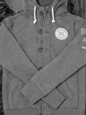 Grå hoodie Hollister med knappar - Snygg grå hoodie från Hollister med knappar framtill, vit logga på bröstet och tryck på ärmen. Utrustad med huva och vita dragsnören. Mjuk bomullsmix och två fickor framtill. Perfekt för en avslappnad och trendig look. Passar runt 165-170. 