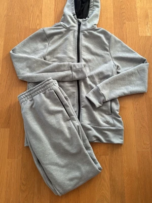 Grått mjukisset med hoodie - Säljer ett grått mjukisset med zip-hoodie och matchande byxor. Hoodien har dragkedja, fickor och huva med svart innerfoder. Byxorna har resår i midjan och sidofickor. Perfekt för chill dagar hemma eller när du vill vara bekväm. Tröja storlek S och byxor M