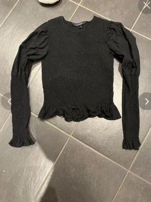 Svart långärmad topp från KappAhl - Trendig svart långärmad topp från KappAhl med smockad kropp och volangdetaljer vid ärmslut och nederkant. Puffiga axlar ger en snygg siluett och toppen har rund halsringning. Perfekt att styla med jeans eller kjol.