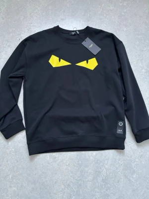 Svart sweatshirt  - Cool svart sweatshirt  med ikoniska gula ögon framtill. Tröjan har rund hals, ribbade muddar vid ärmslut och nederkant samt en avslappnad passform. Perfekt för dig som vill sticka ut med en unik detalj. Materialet känns mjukt och bekvämt.