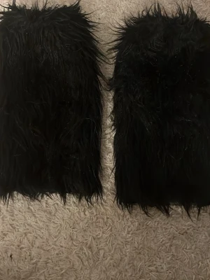 Svarta fluffiga benvärmare cosplay - Ett par svarta, långa och fluffiga benvärmare perfekta för maskerad eller cosplay. Tillverkade i syntetiskt pälsmaterial som ger en cool och iögonfallande look. Passar till rave, festival eller temafest.