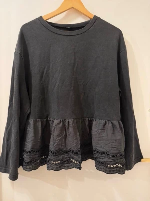 Svart grå urtvättat färg blus med volang och spetsdetaljer - En svart grå  blus med långärmad design och rund hals. Nederdelen har en volang med dekorativa spetsdetaljer som ger en cool och unik look. Perfekt för dig som gillar att sticka ut med snygga detaljer. Materialet känns som bomull upptill och syntet i volangen.