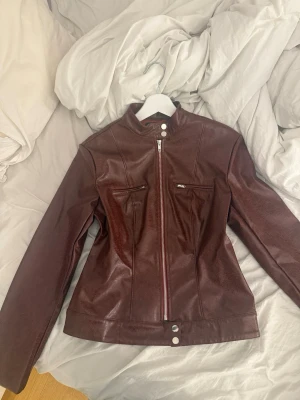 Mörkbrun skinnjacka med ståkrage - Snygg röd skinnjacka med ståkrage och dragkedja framtill. Två bröstfickor med dragkedja och markerad midja för en schysst siluett. Perfekt för dig som vill ha en stilren och tidlös look. Biker jacka, använd två gånger, köpt för runt 450 från shein, kvaliteten e jätte bra, säljer då jag inte använder den 