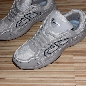 Grå sneakers från Dior - Snygga grå sneakers från Dior med mesh och syntetpaneler. Skorna har en sportig design med svarta och vita detaljer längs sidorna och en tydlig Dior-logga på plösen. Klassisk snörning och vadderad häl för extra komfort. Perfekta för en trendig streetwear-look.