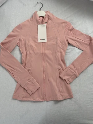 Rosa zip-jacka från Lululemon - Snygg rosa träningsjacka från Lululemon med figurnära passform, hel dragkedja framtill och två fickor med dragkedja. Tillverkad i ett stretchigt och mjukt funktionsmaterial som passar perfekt till träning eller som ett sportigt lager. Liten logga på dragkedjan. Helt oanväbd med prislapp eftersom att ja fick de i present och den var för liten. STORLEK 4