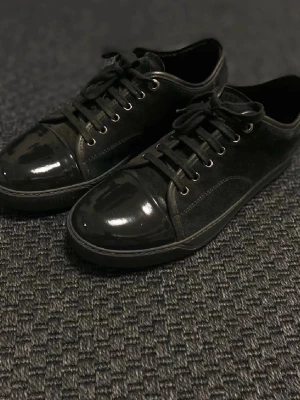 Byte - Stilrena svarta sneakers med glansiga lackdetaljer på tån och klassisk snörning. Ovandelen är i en mix av mocka och textil, vilket ger en snygg kontrast till den blanka tåkappan. Perfekta för dig som gillar en clean och modern look.