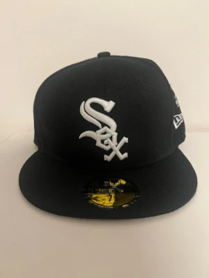Svart White Sox keps från New Era - Säljer en svart 59FIFTY keps från New Era med White Sox-logga broderad i vitt framtill. Klassisk flat brim och strukturerad passform. Kepsen har New Era-logga på sidan och är tillverkad i ett slitstarkt material.