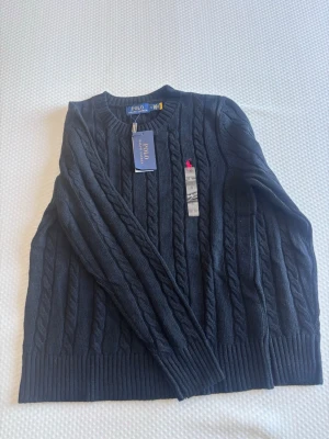 Stickad tröja polo Ralph lauren - Snygg mörkblå kabelstickad tröja från Polo Ralph Lauren. Tröjan har rund halsringning, ribbade muddar och klassisk kabelstickning. Tillverkad i mjuk bomull och har en liten rosa logga broderad på bröstet. Perfekt för en stilren och avslappnad look. Helt ny polo passar ej mig skriv vid frågor 