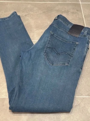 Blå jeans från Nudie Jeans - Säljer ett par klassiska blå jeans från Nudie Jeans med rak passform och fem fickor. Jeansen har en mörkblå tvätt och är tillverkade i bomull. Snygg detalj med Nudie-loggan på bakfickan och läderpatch i midjan.