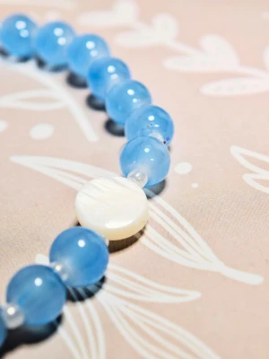 Blått pärlarmband med vit detalj - Snyggt armband med runda, ljusblå glaspärlor och en större, platt vit pärla som accent. Armbandet har elastisk tråd och passar perfekt för dig som gillar färgglada accessoarer. Elastiskt armband av hög kvalitet, 18 cm långt.