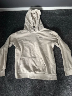 Beige hoodie från Hollister - Snygg beige hoodie från Hollister med klassisk huva och stor magficka. Diskret Hollister-logga på bröstet. Tillverkad i mjukt bomullsmaterial som känns skönt mot huden. Perfekt för en avslappnad och stilren look.