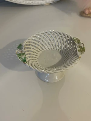 Vit flätad porslinsskål på fot - Unik vit skål i porslin med flätad design och två handtag dekorerade med gröna detaljer. Skålen står på en rund fot och har ett öppet, luftigt mönster. Perfekt som dekoration eller för småsaker.