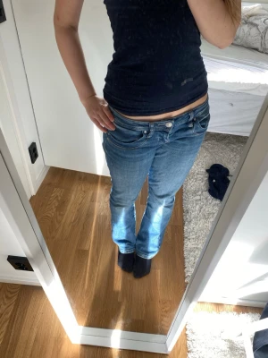 Blå bootcut jeans med låg midja - Säljer ett par klassiska blå bootcut jeans med låg midja och fem fickor. Jeansen har en snygg tvätt och subtila slitningar för en avslappnad look. Tillverkade i mjuk denim med stretch för extra komfort. Perfekta för dig som gillar en tidlös och trendig stil.