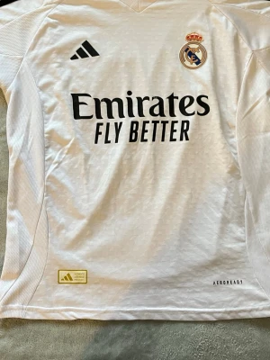 Vit Real Madrid matchtröja Adidas - Snygg vit Real Madrid matchtröja från Adidas med klubbmärke och sponsortryck på bröstet. Tröjan har svarta detaljer, rund hals och är tillverkad i lätt, ventilerande material med AEROREADY-teknologi. Perfekt för fotbollsträning eller att bära till vardagarna.