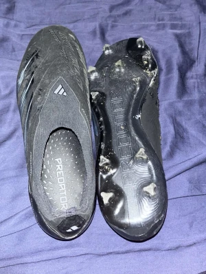 Adidas Predator Elite svarta fotbollsskor - Säljer ett par svarta Adidas Predator Elite fotbollsskor med strömlinjeformad design och utan snörning. Skorna har texten 'PREDATOR' på innersulan och yttersulan samt 'HYBRIDTOUCH 2.0' på sidan. Utrustade med fasta dobbar för bra grepp på planen.