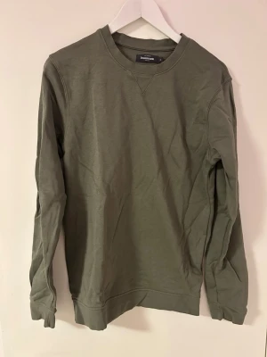 Olivgrön sweatshirt från dressmann - En stilren olivgrön sweatshirt från Dressmann med rund halsringning och långa ärmar. Tröjan är gjord i mjuk bomull och har ribbade muddar vid ärmslut och nederkant. Perfekt för en avslappnad look.