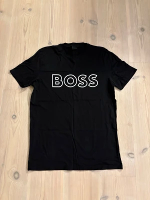  BOSS t-shirt - Snygg svart t-shirt från BOSS med stort vitt logotryck på bröstet. Klassisk rund halsringning och korta ärmar. Perfekt basplagg med modern och stilren design, tillverkad i mjuk bomull för skön känsla.