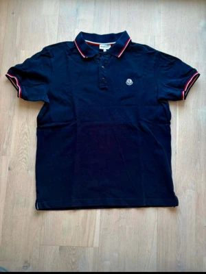 Marinblå pikétröja från Moncler - Snygg marinblå pikétröja från Moncler med klassisk krage och knäppning framtill. Tröjan har röda och vita ränder på krage och ärmslut samt Moncler-logga på bröstet. Perfekt för en stilren och sportig look.