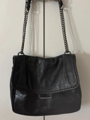 Svart handväska med kedjerem - Snygg svart handväska i konstläder med lock och dekorativa sömmar. Väskan har en silverfärgad kedjerem och ett innerfack med dragkedja. Perfekt storlek för det viktigaste och ger en cool vibe till din outfit.