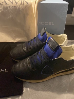 Philippe Model svarta sneakers i skinn - Snygga svarta sneakers från Philippe Model med blåa detaljer och vit insida. Skorna har klassisk snörning, rund tå och en beige sula. Tillverkade i skinn med textil på plösen och sidan. Perfekta för dig som gillar stilrena men ändå unika sneakers. Skorna är även knappast använda, köpte de i början av november sedan har jag använt de ett väldigt få par gånger. Du får även med Lanvin dustbag och deras egna dustbag samt box. För ytterligare bilder och funderingar får du gärna höra av dig. (: