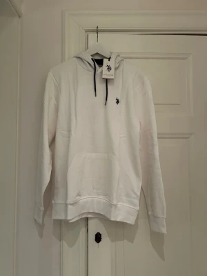 Vit hoodie från U.S. Polo Assn. - Snygg vit hoodie från U.S. Polo Assn. med svart dragsko i huvan och klassisk logga broderad på bröstet. Hoodien har en stor magficka och ribbade muddar vid ärmar och nederkant. Tillverkad i mjukt bomullsmaterial för skön känsla.