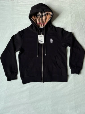 Svart Burberry hoodie med dragkedja - Svart hoodie från Burberry med dragkedja framtill och klassiskt rutigt foder i huvan. Broderad vit TB-logga på bröstet och två fickor fram. Snygg och stilren design som passar perfekt till streetwear-looken.