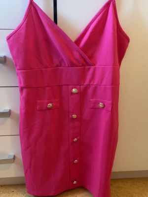 Rosa kort klänning med knappar - Snygg rosa kort klänning med omlott upptill och dekorativa silverknappar framtill. Klänningen har smala axelband och två fickdetaljer framtill. Perfekt för sommarens alla tillfällen.