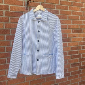 Randig overshirt från John Henric - Snygg randig overshirt från John Henric i blått och vitt. Jackan har klassisk krage, stora svarta knappar och två rejäla fickor framtill. Tillverkad i luftig bomull, perfekt för lager-på-lager. Långärmad och stilren med relaxed vibe.