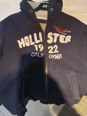 Mörkblå Hollister hoodie med dragkedja - Mörkblå hoodie från Hollister med broderad logga och text på bröstet. Tröjan har huva med snören, dragkedja framtill och två stora fickor. Materialet är mjukt och bekvämt, perfekt för chill dagar. Ikonisk Hollister-fågel på bröstet och detaljer i vitt och rött.