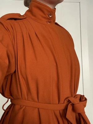 Rodebjer - Roströd trenchcoat med knytskärp - Vacker roströd trenchcoat med hög krage och dubbla knappar. Knytskärp i midjan och axelklaffar ger en klassisk look. 