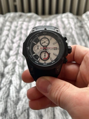 Daniel Steiger sportig kronografklocka - Säljer en cool kronografklocka från Daniel Steiger med svart boett och armband i syntetmaterial. Urtavlan är vit med flera små visare och röda detaljer. Klockan har robust design och tydliga siffror, perfekt för dig som gillar sportiga accessoarer. Ny pris 1500. 10 ATM vattentät 
