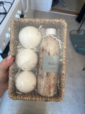 Presentkit med badbomber och badsalt - Snyggt presentkit med tre vita badbomber och en flaska badsalt i en naturlig korg. Badbomberna har små bruna inslag och badsaltet kommer i en genomskinlig plastflaska med etikett. Perfekt för avkoppling och spa-känsla hemma.