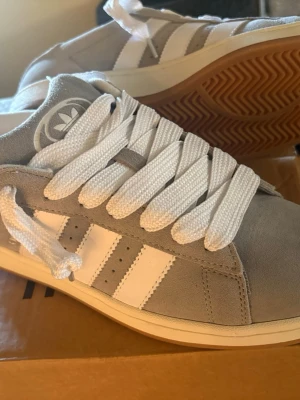 Adidas campus - Snygga sneakers från Adidas i grå mocka med klassiska vita ränder på sidan och vit sula. Skorna har breda vita skosnören och en brun yttersula med mönster. Perfekta för dig som gillar stilrena och sportiga skor. Som ny!!