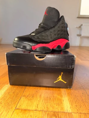 Air Jordan 13 Retro Black/Red - Säljer ett par Air Jordan 13 Retro sneakers i svart och rött. Skorna har en hög siluett, mesh och lädermaterial, samt ikoniska Jumpman-loggan på plösen. Sulan är robust och designen är inspirerad av basket. Perfekt för dig som vill sticka ut med klassiska Jordans.