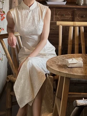 Vit långklänning med broderad spets - Elegant vit långklänning med hög krage och ärmlös design. Klänningen har vackra broderade spetsdetaljer och en slits framtill. Materialet är troligtvis satin eller silke, vilket ger en lyxig känsla och fin struktur.