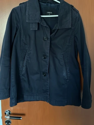 Mörkblå trenchcoat från Lindex - Stilren svart trenchcoat från Lindex med klassisk krage, knäppning framtill och två snedställda fickor. Jackan har axelremmar och är tillverkad i ett lätt material som passar perfekt för lager-på-lager. Snygg och enkel att matcha med olika outfits.