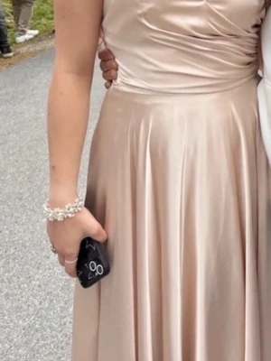 Champagnefärgad långklänning i satin - Elegant långklänning i champagnefärgad satin med draperad överdel och utsvängd kjol. Klänningen har en klassisk och stilren design som ger ett lyxigt intryck. Perfekt för speciella tillfällen och högtider.160-178cm