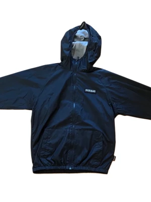 Svart vindjacka från Napapijri - Snygg svart vindjacka från Napapijri med huva och dragkedja framtill. Jackan har två stora fickor, elastisk nederkant och är tillverkad i ett lätt, vattenavvisande material. Perfekt för blåsiga dagar och enkel att matcha med olika outfits.