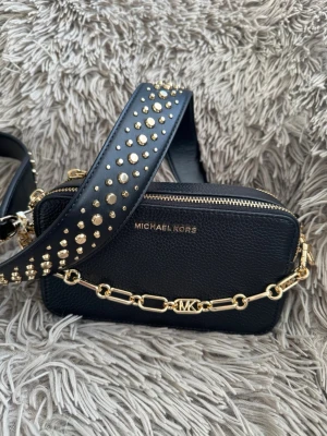 Svart Michael Kors handväska med nitar - Snygg svart handväska från Michael Kors i läder med guldfärgade detaljer. Väskan har en bred axelrem dekorerad med guldnitar och en kedja framtill med MK-logga. Perfekt storlek för det viktigaste och stängs med dragkedja.